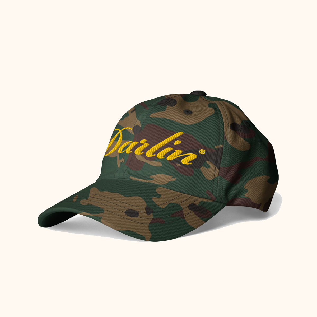 Angled view of DARLIN® original script camouflage hat with yellow embroidery on a warm ivory background