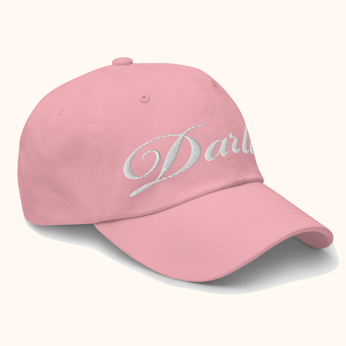 Angled view of pink DARLIN® script hat with white embroidery on a warm ivory background