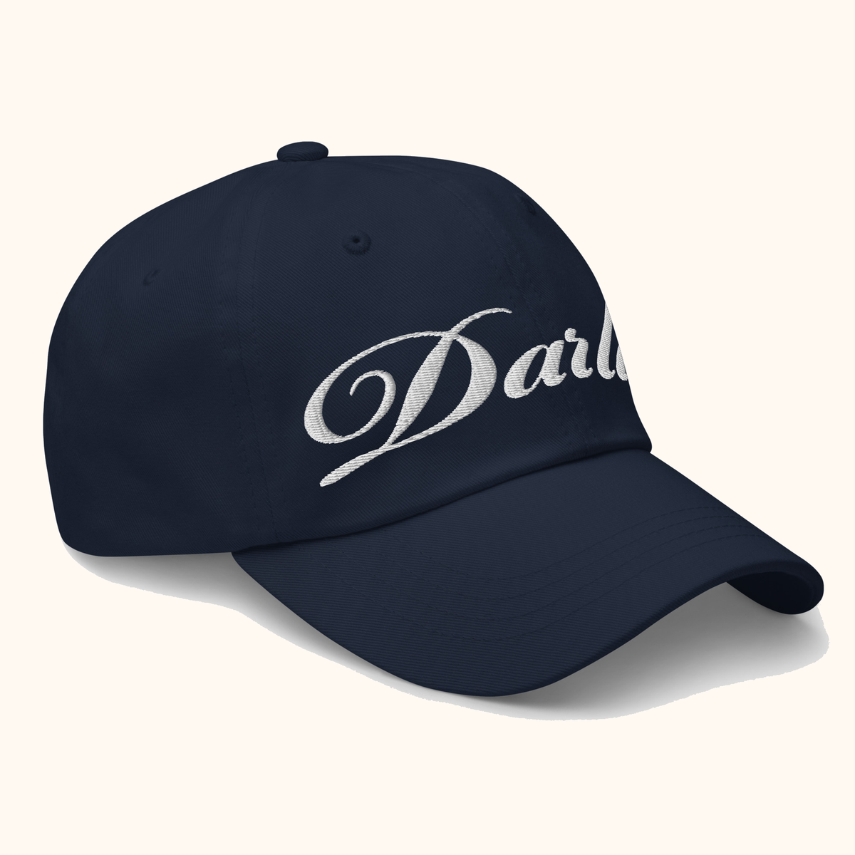 Angled view of DARLIN original script navy hat with white embroidery on a warm ivory background