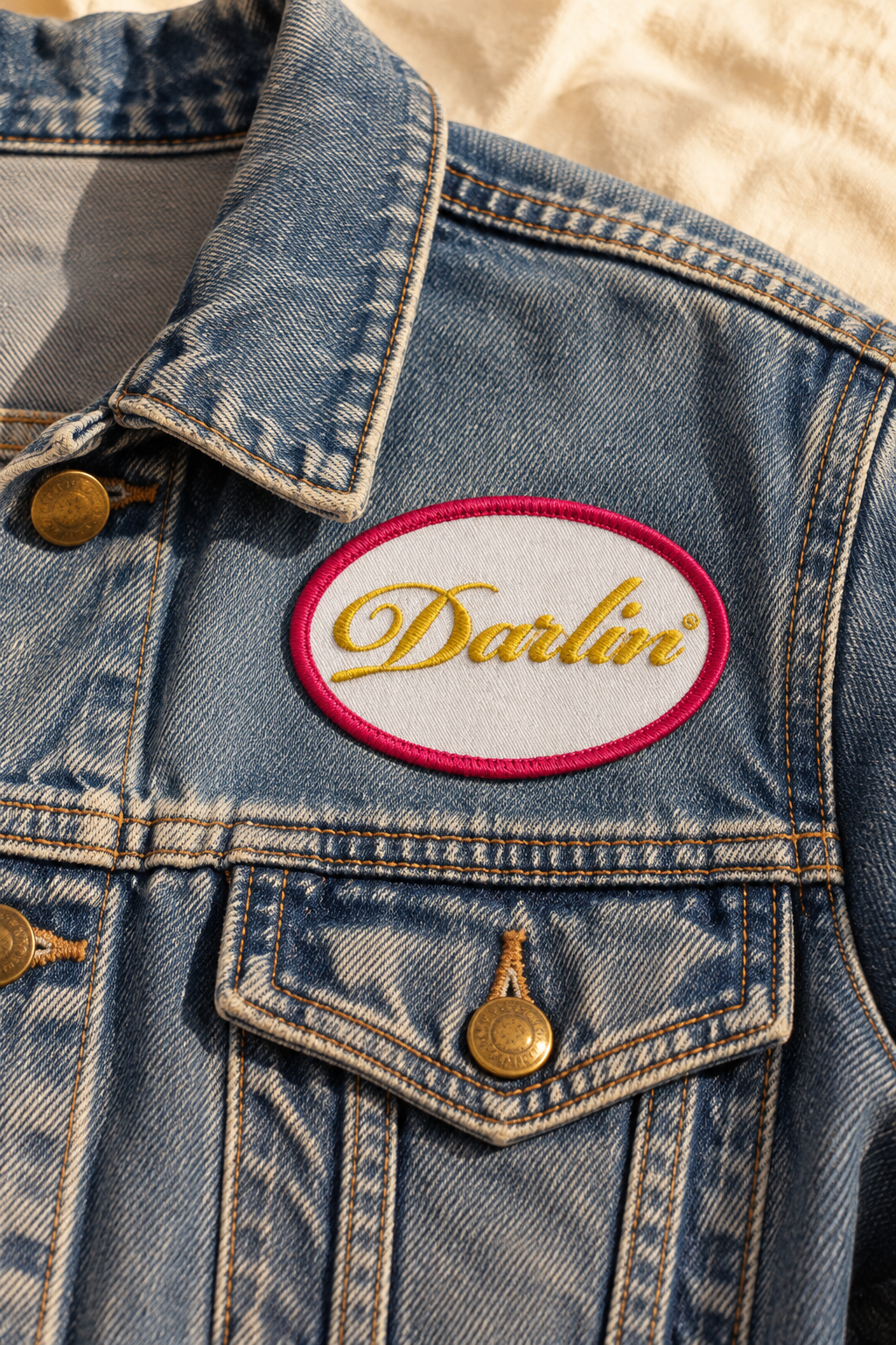 Close-up of a white oval DARLIN® patch with gold script embroidery and a fuchsia border on a vintage blue denim jacket.